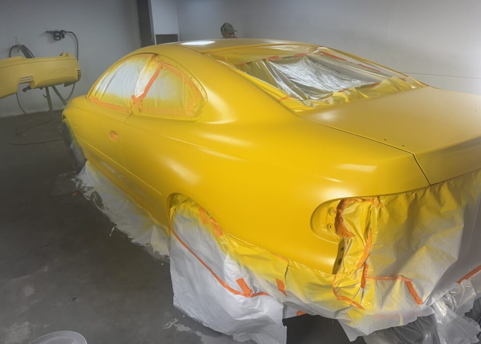 Side view of a yellow Pontiac GTO being professionally painted inside a controlled spray booth at D&R Auto Body & Paint.