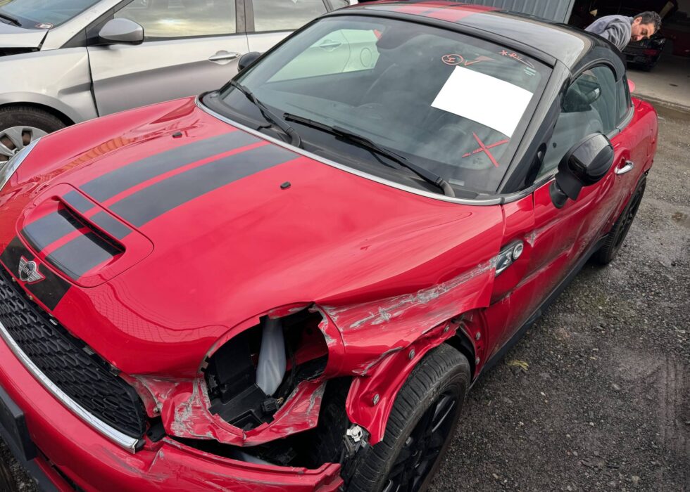 Red Mini Cooper with front‑end collision damage, missing headlight, and crumpled fender at D & R Autobody & Paint repair shop.