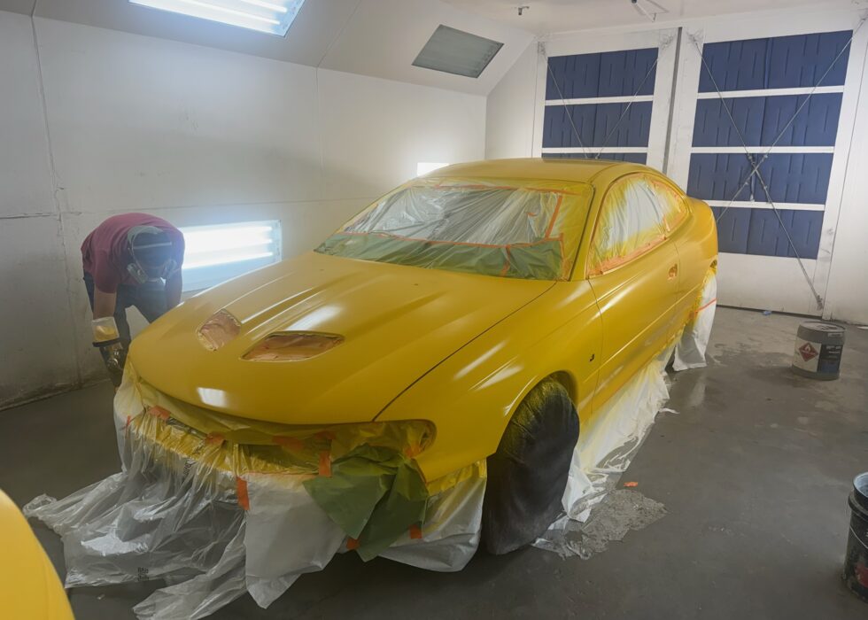 A yellow Pontiac GTO inside a professional spray booth at D&R Auto Body & Paint, masked off with paper and tape for a custom paint job.