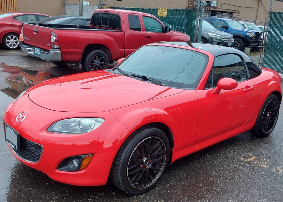 Red Mazda MX-5 Miata with a black retractable hardtop and custom black wheels parked at D&R Auto Body & Paint.