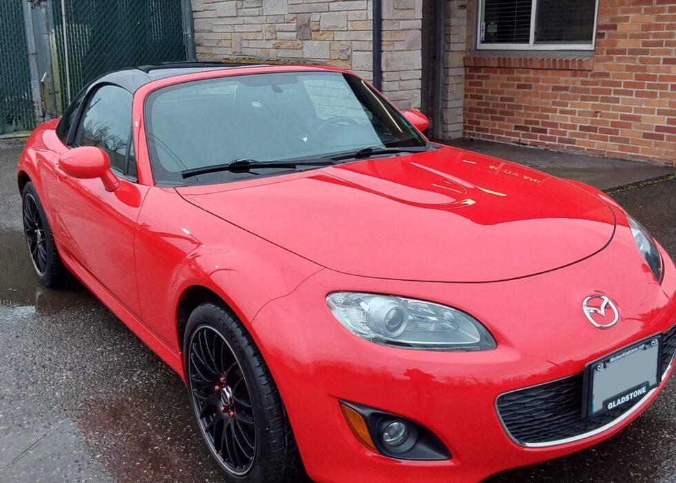 Red Mazda MX-5 Miata with a black retractable hardtop and custom black wheels parked in front of the D&R Auto Body & Paint shop.